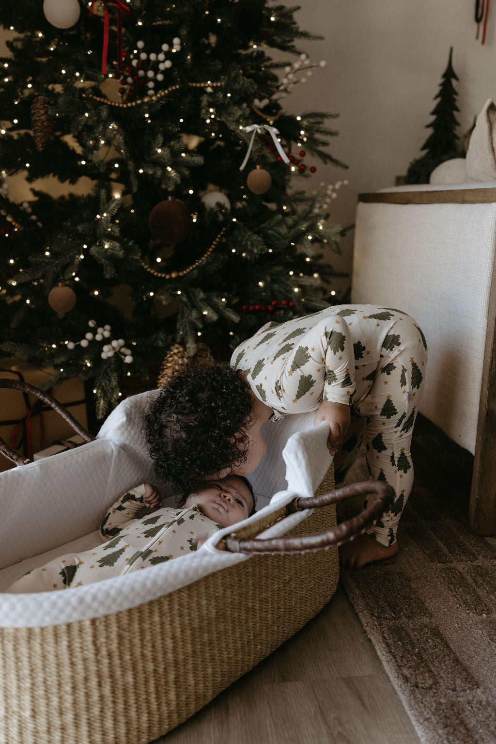 Child in a crib next to a decorated Christmas tree in a cozy room.